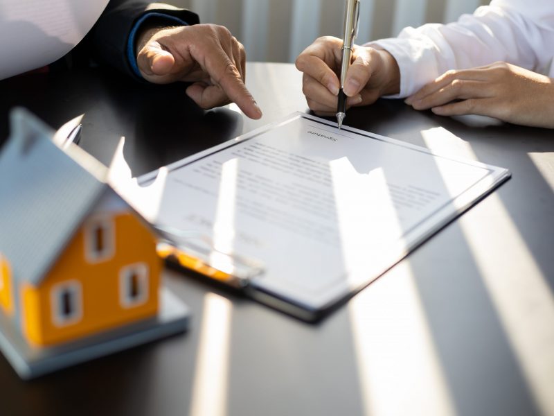 Businessman in suit in his office showing home insurance policy and pointing with a pen where the policyholder must to sign. Insurance agent consulting real estate insurance detail to customer.